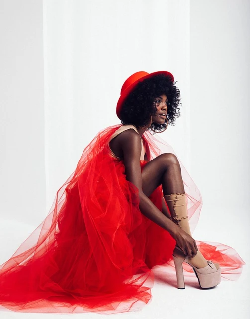 Woman in red tulle dress and matching hat adjusting high-heeled shoe in a photo studio. Beauty and fashion photography.