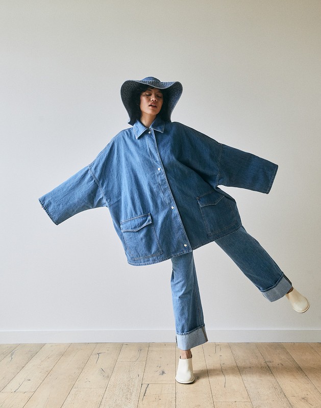 Model in oversized denim jacket, matching hat and jeans, striking a dynamic pose against a white backdrop on a wooden floor.