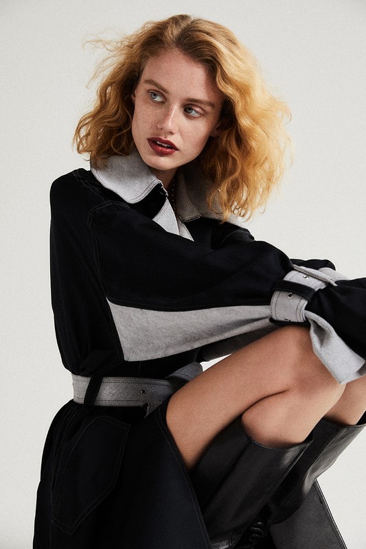 Model with blonde curly hair and dark lipstick wearing a black and grey belted coat against a white background
