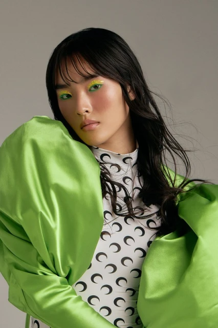 Portrait of a stylish woman with bright green eye makeup wearing a patterned top and a lime green jacket in a photo studio.