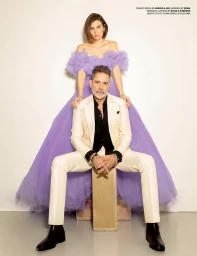 Woman in an off-the-shoulder purple tulle gown standing behind a seated man in a white suit and black shirt against a plain background.