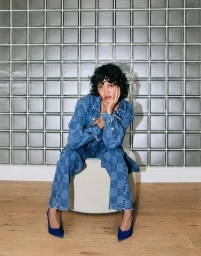 Woman with dark curly hair wearing a blue patchwork denim jacket and trousers seated on a white stool against a glass block wall