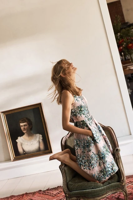 A woman in a floral dress sits on a chair in a sunlit room, next to an antique portrait and a patterned red rug.