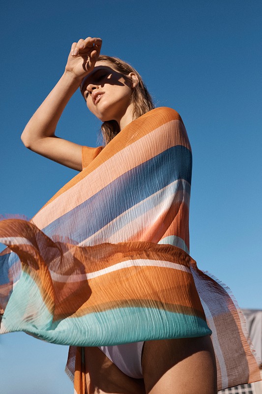 Woman wearing colorful striped shawl poses against clear blue sky in outdoor setting on a sunny day. Fashion and lifestyle photography.