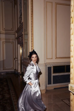 Elegant adult woman wearing a silver dress with a trench coat and a bow in her hair, walking inside a building with classic architecture.