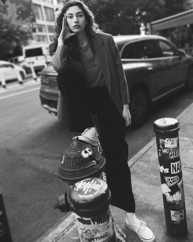 Monochrome outdoor fashion shot of a young woman posing on a fire hydrant, wearing an oversized blazer and wide-leg pants, with a car in the background.