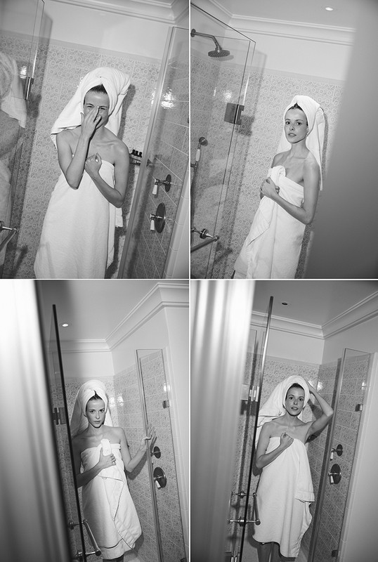 Monochrome portrait of a woman in a shower covered in a towel, with a towel on her head, standing near shower fixtures and glass door.