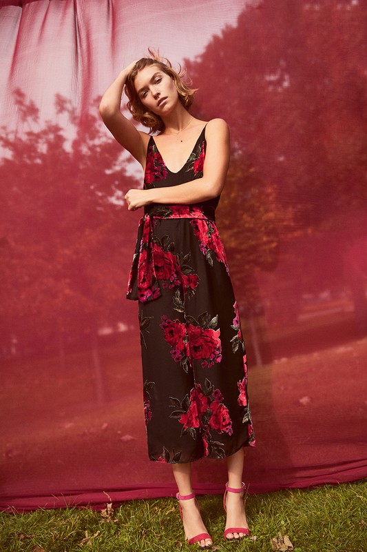 A fair-skinned woman in a black floral dress and pink heels poses in front of a red and green background, holding her hand on her head.
