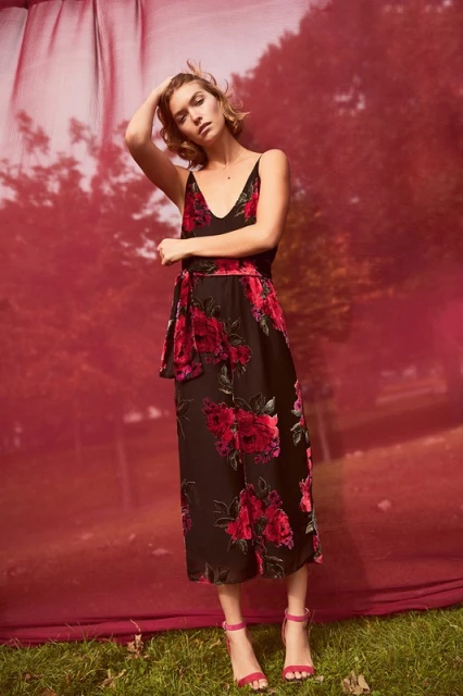 A fair-skinned woman in a black floral dress and pink heels poses in front of a red and green background, holding her hand on her head.