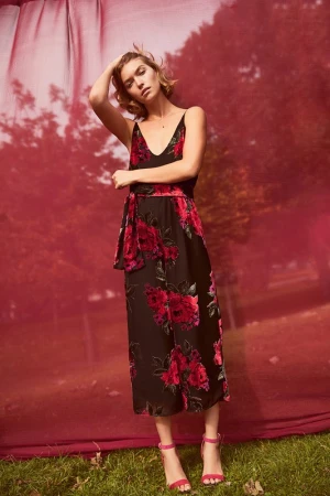 A fair-skinned woman in a black floral dress and pink heels poses in front of a red and green background, holding her hand on her head.