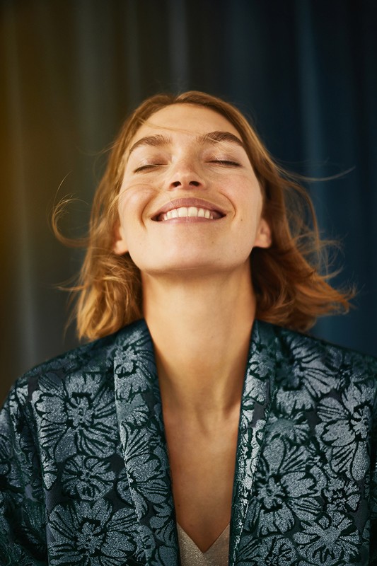 Smiling adult woman with closed eyes, auburn hair, and floral-patterned top, standing in front of a textured blue background.