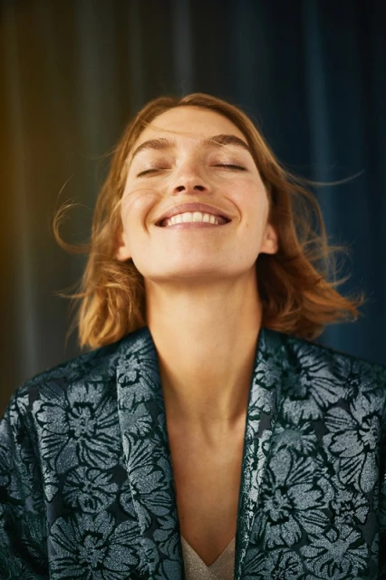 Smiling adult woman with closed eyes, auburn hair, and floral-patterned top, standing in front of a textured blue background.