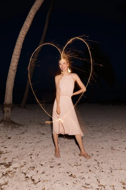 Adult woman in a dress poses with a sparkler in the shape of a heart on a beach at night with palm trees in the background.