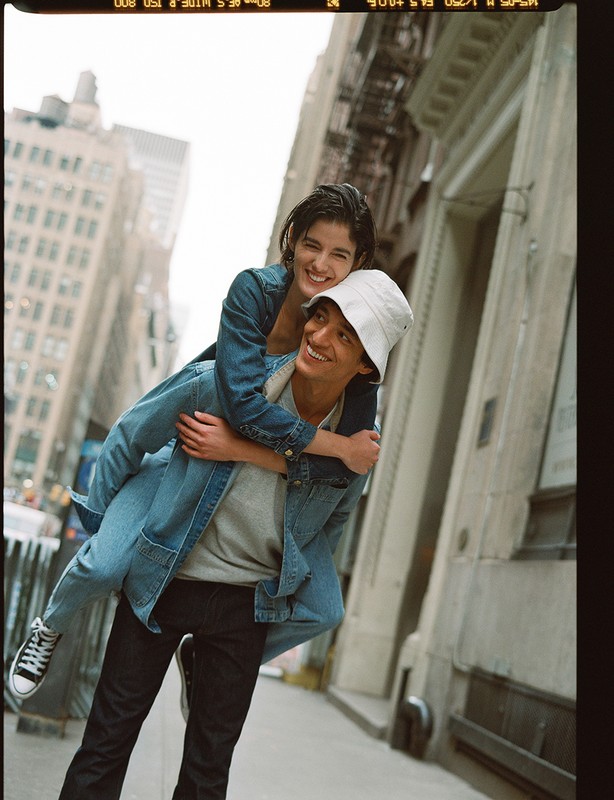 Smiling couple enjoying piggyback ride on city street with tall buildings, blue denim jackets, and youthful casual fashion.