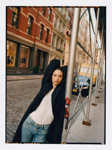 Woman in a black coat poses against scaffolding on a city street with brick buildings and parked cars under natural light.