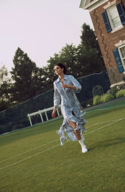 Woman in patterned dress runs across a grassy lawn toward a brick building on a sunny day, showcasing a fashion editorial.
