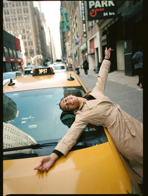 Woman in trench coat laying on taxi in urban setting. Smiling adult female with arms outstretched on yellow taxi cab in city.