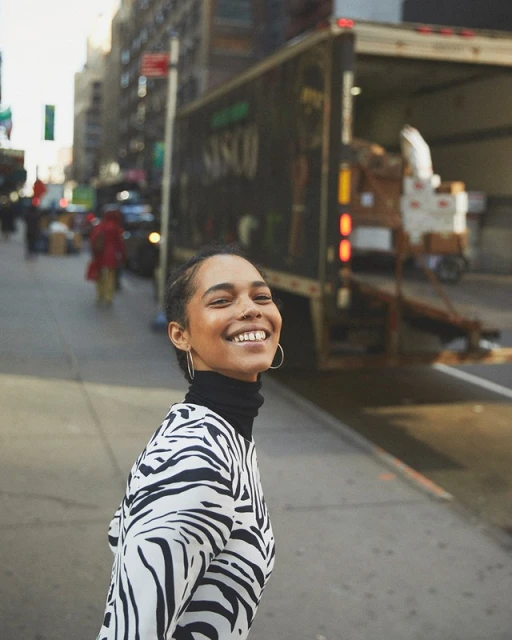 Smiling woman in zebra-print top on a city sidewalk with a commercial truck, buildings, and pedestrians in the background.