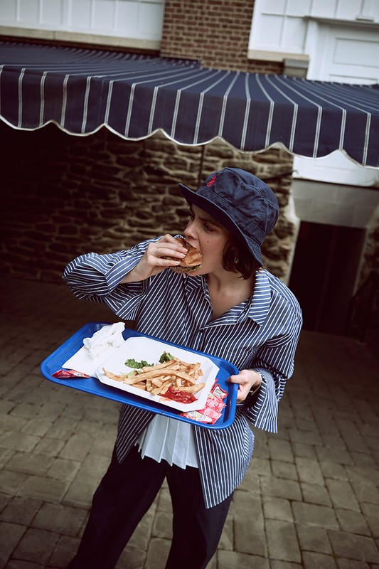 Adult female in a striped shirt and bucket hat is eating a burger while holding a tray of French fries in an outdoor setting.
