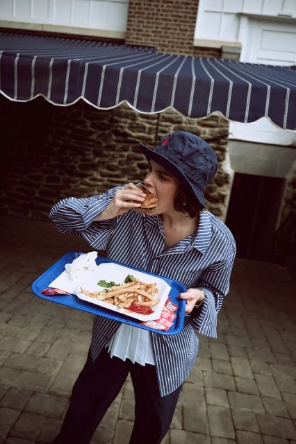 Adult female in a striped shirt and bucket hat is eating a burger while holding a tray of French fries in an outdoor setting.