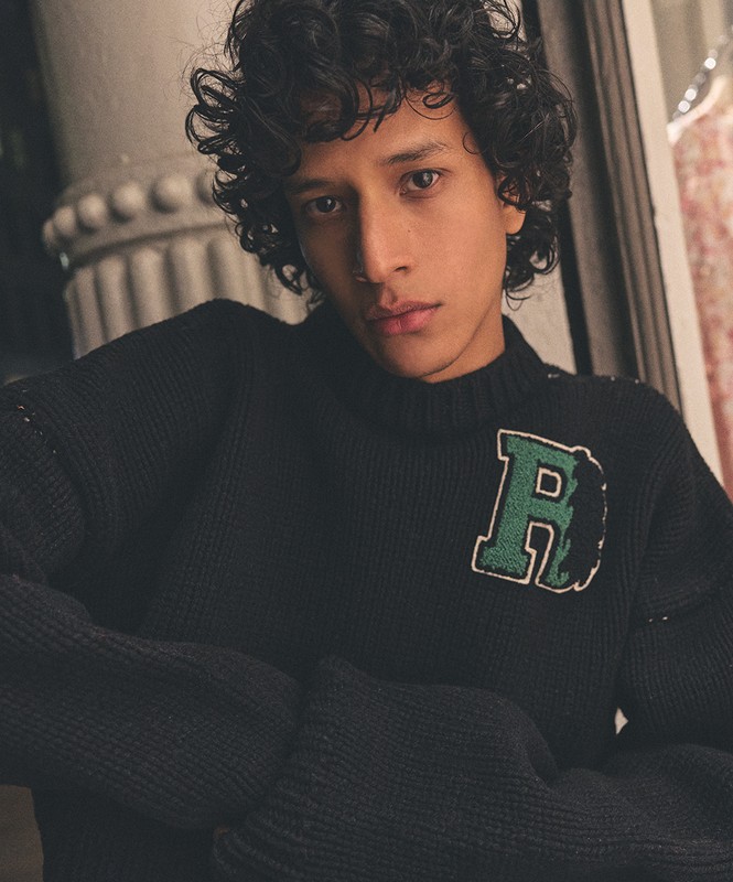 Portrait of a handsome curly-haired man wearing a knit sweater with an initial. Indoor setting, soft lighting, medium shot, portrait orientation.