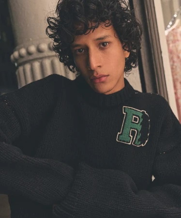 Portrait of a handsome curly-haired man wearing a knit sweater with an initial. Indoor setting, soft lighting, medium shot, portrait orientation.