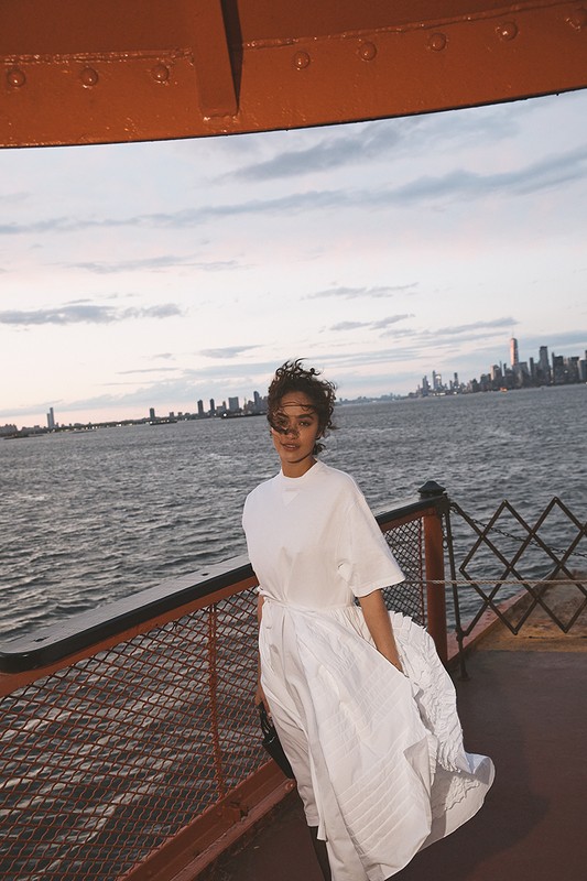 A young woman wearing a white dress standing on a ferry with the New York City skyline and blue sky in the background.