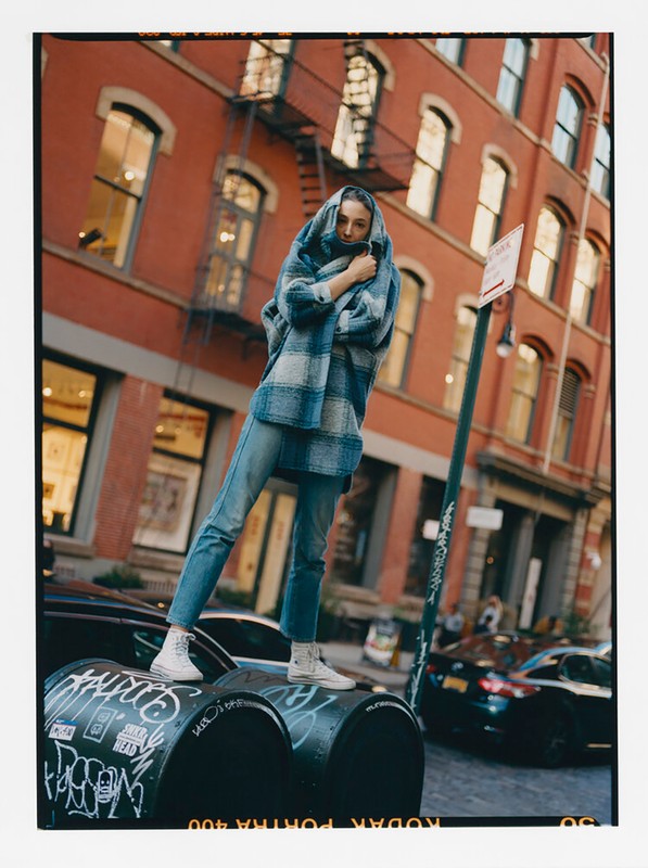 Outdoor fashion shot of a young adult female model wearing a blue plaid hooded coat and jeans, standing on black metal containers.