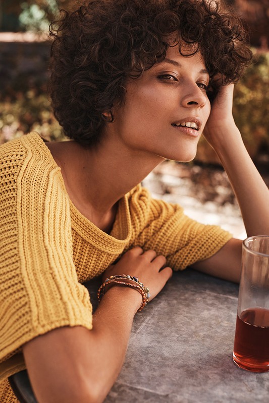 Woman with short curly hair wearing a mustard yellow knitted top seated outdoors near a glass of red beverage.