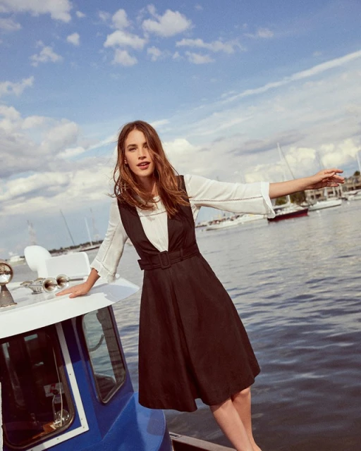 Woman in vintage-style black dress poses on a blue boat under a sunny sky with other sailboats on the water in the background.