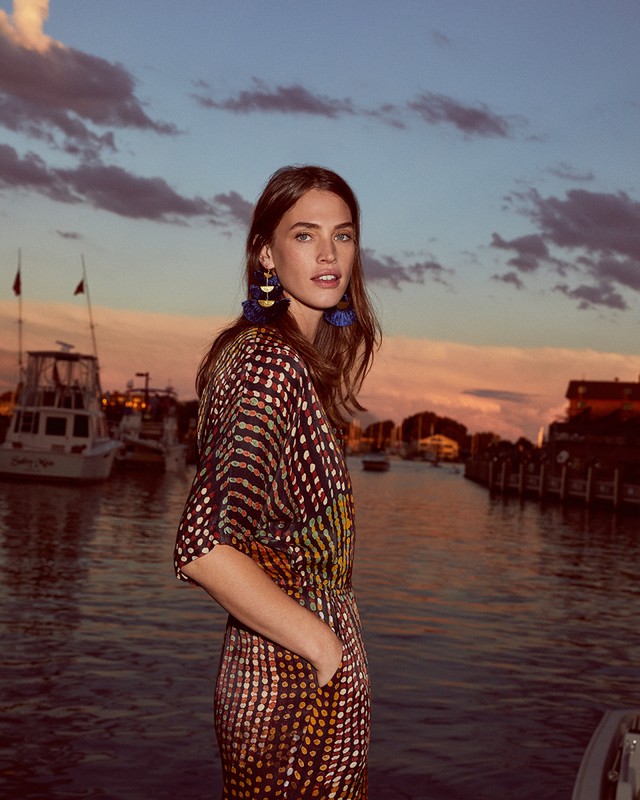 A fashionable woman stands near a pier at sunset in a polka dot dress with blue tassel earrings near boats in the background.