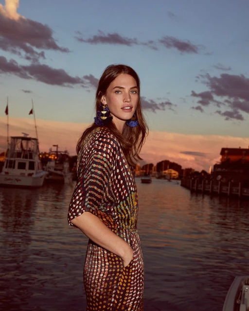 A fashionable woman stands near a pier at sunset in a polka dot dress with blue tassel earrings near boats in the background.