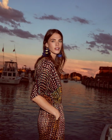 A fashionable woman stands near a pier at sunset in a polka dot dress with blue tassel earrings near boats in the background.