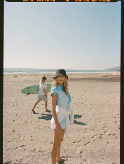 Woman in bucket hat poses on beach with man carrying surfboard in background under clear sky on sunny day. Coastal scene.