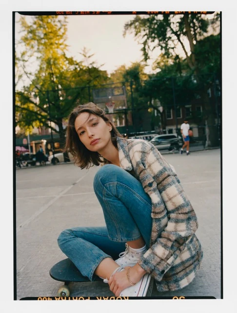 A woman with short brown hair sits on a skateboard in a park wearing jeans, a plaid jacket, and sneakers in an eye-level shot.
