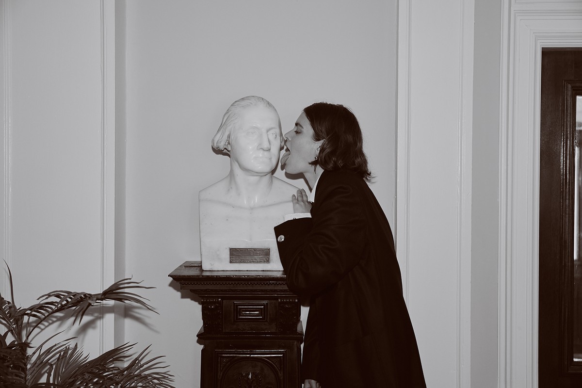 A woman touches her tongue to a George Washington bust in a black-and-white photo with a plant, door, and wall in the background.