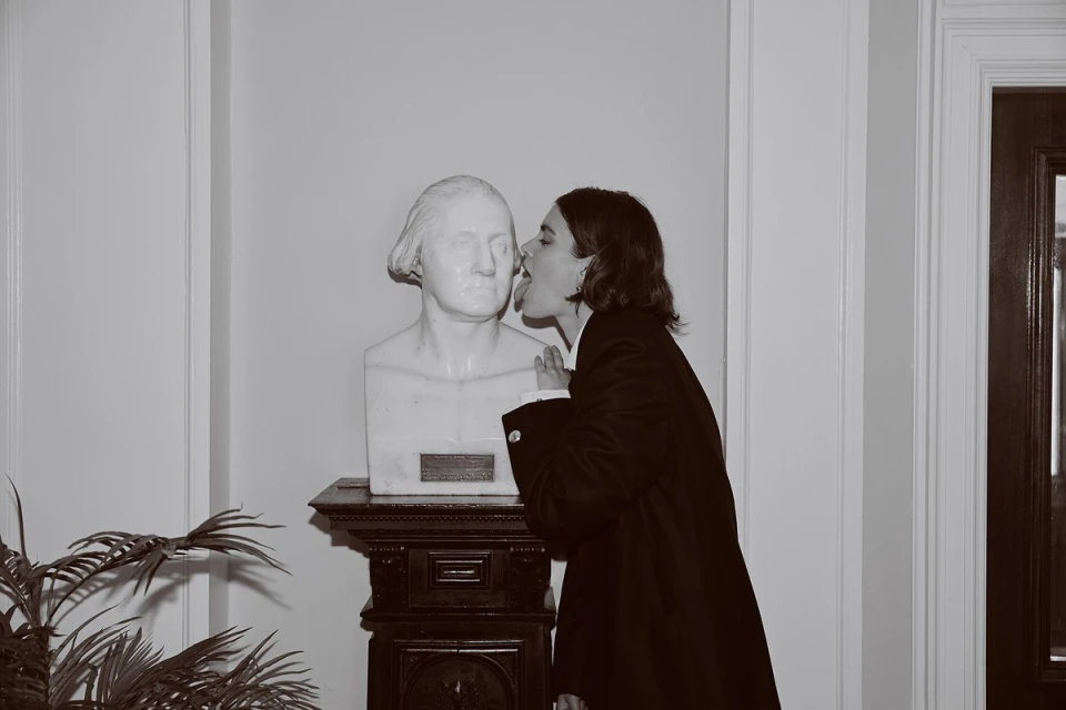 A woman touches her tongue to a George Washington bust in a black-and-white photo with a plant, door, and wall in the background.