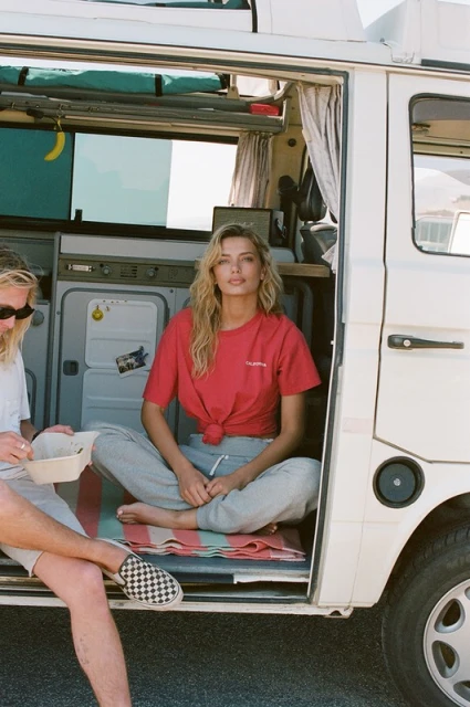 A woman sits in the doorway of a white camper van wearing a red t-shirt and grey sweatpants, with a man sitting beside her.