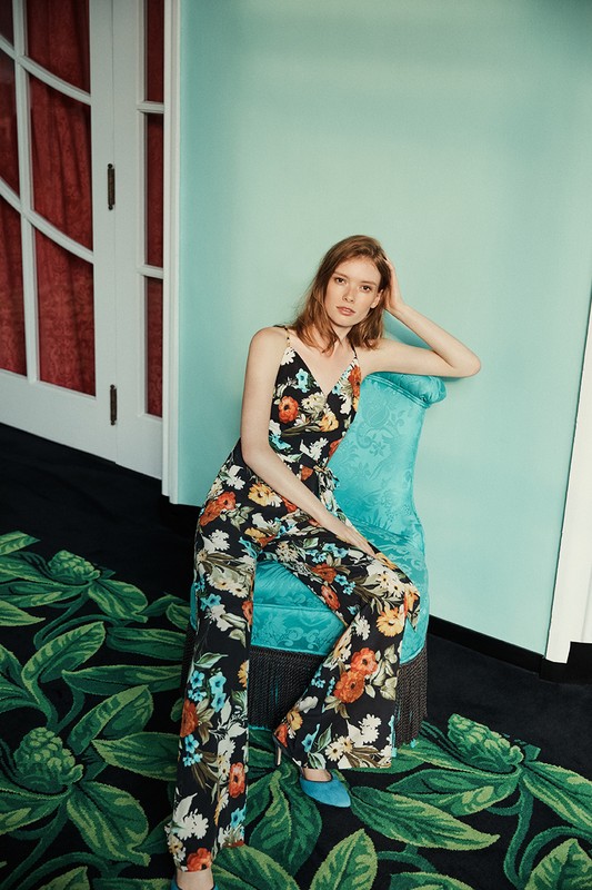 Elegant woman in a floral-patterned jumpsuit sits on a turquoise chair against a light blue wall, complemented by a patterned rug.