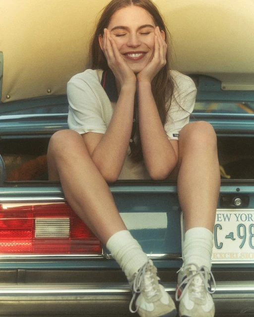 Smiling woman in a white t-shirt sitting on the back of a vintage car with New York license plate in an eye-level shot.
