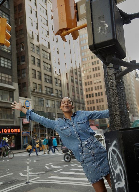 Smiling woman in denim outfit poses with arms outstretched next to a traffic light in a city street with pedestrians and buildings.