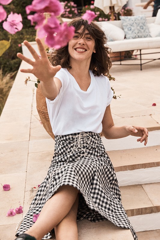 A woman in a white t-shirt and black and white checkered skirt smiles while sitting outside surrounded by pink flowers and greenery.