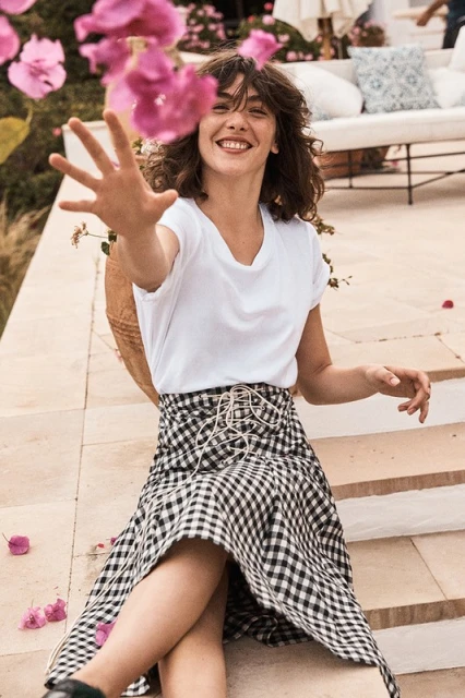A woman in a white t-shirt and black and white checkered skirt smiles while sitting outside surrounded by pink flowers and greenery.