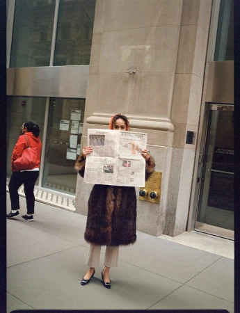 Woman holding up a newspaper in front of her face, wearing a fur coat on a city sidewalk photographed by Landon Mcmahon