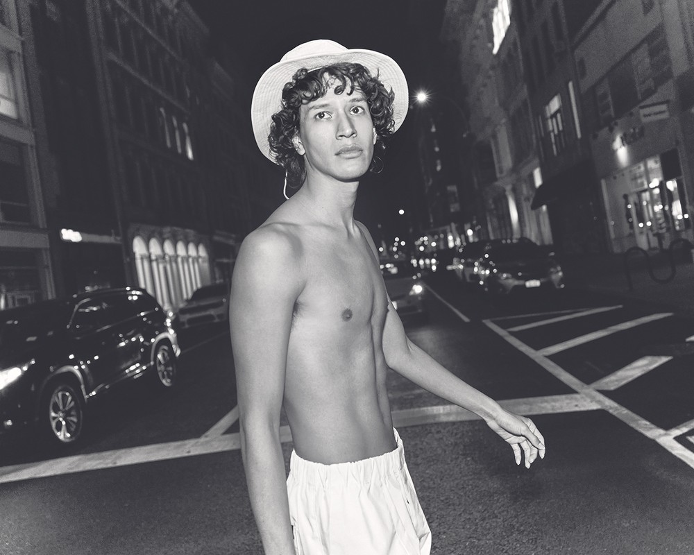 Black and white image of a shirtless man with a hat standing on a crosswalk at night in a city with car and building background.