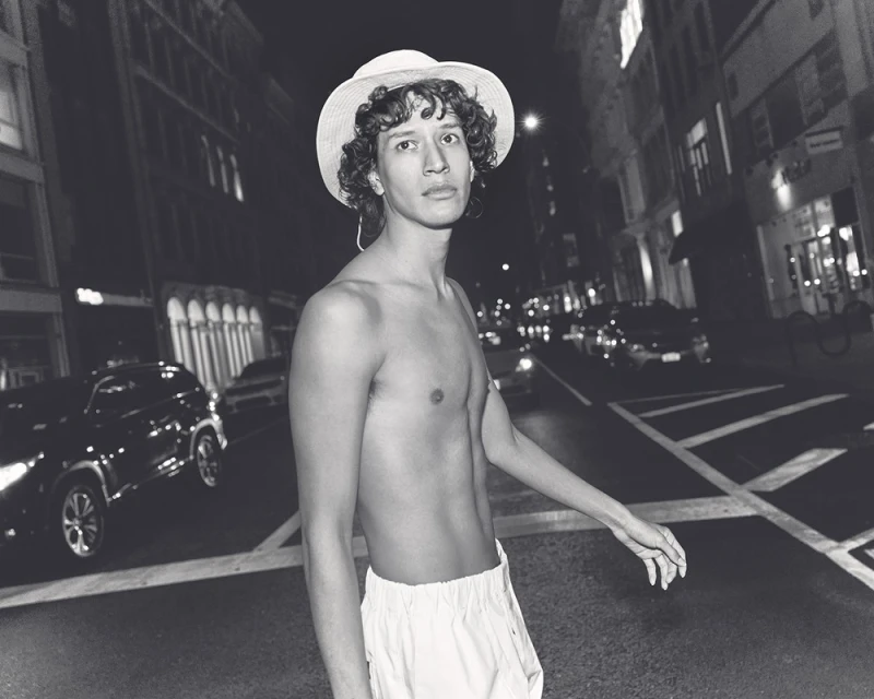 Black and white image of a shirtless man with a hat standing on a crosswalk at night in a city with car and building background.