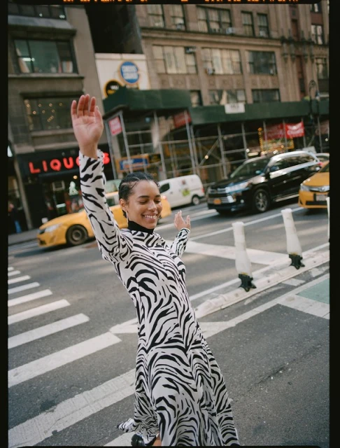 Young woman in zebra-print dress joyfully poses mid-street crossing in New York City, smiling with arms raised, surrounded by city traffic and buildings.