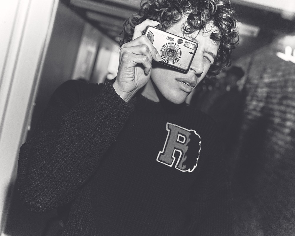 Young man in a sweater holds a compact camera up to his face indoors in a black-and-white photo. He has curly hair and dark clothes.