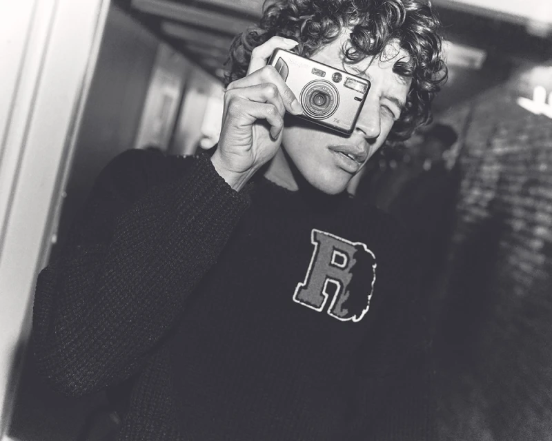 Young man in a sweater holds a compact camera up to his face indoors in a black-and-white photo. He has curly hair and dark clothes.