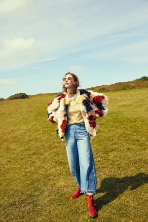 Model wearing a fluffy patchwork faux-fur jacket and blue jeans standing in a grassy field under a blue sky.